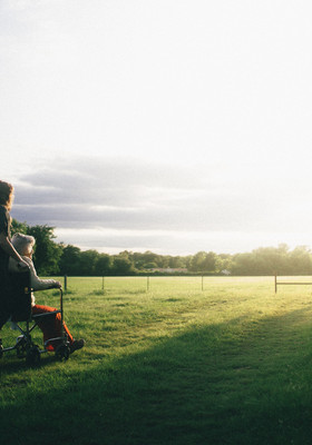Alte Dame in Rollstuhl und Pfleger betrachten die aufgehende Sonne über einem Feld