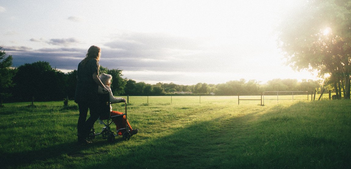 Alte Dame in Rollstuhl und Pfleger betrachten die aufgehende Sonne über einem Feld